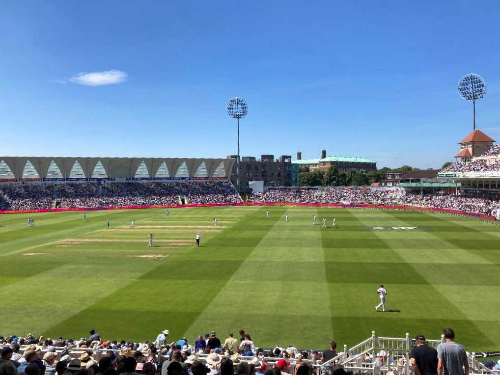 A wide view of a Test match being played in a sunny stadium, taken from behind the spectators